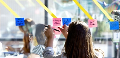 Vrouw die Post-it® Super Sticky Note 76 x 76 mm op een glazen wand plaatst om de vastgelegde taaknotities te delen met teamgenoten of projecten